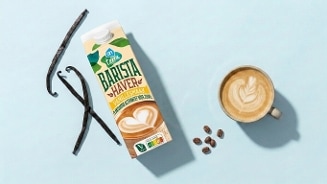 Pak AH Terra Barista Haver Vanillesmaak naast een kopje koffie met latte art, enkele koffiebonen en vanillestokjes op een lichtblauwe achtergrond.
