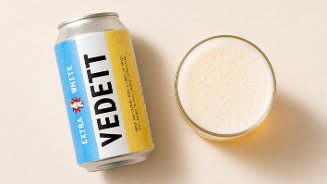 Blik Vedett Extra White bier naast een glas gevuld met witbier op een lichtgekleurde ondergrond.