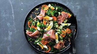 Salade met gesneden wortel, ei, veldbonen, boerenkool, gerookte ham en zaden op een zwart bord met bestek