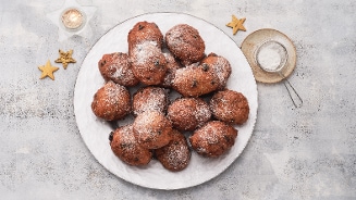Een schaal met oliebollen bestrooid met poedersuiker, omringd door stervormige koekjes, een zeefje met poedersuiker en een brandend waxinelichtje op een grijze achtergrond.