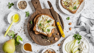 Toast met roomkaas, plakjes peer en noten op een houten snijplank, omringd door verse ingrediënten zoals amandelen, honing en een hele peer.