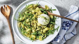 Pastasalade met schelpvormige pasta, rucola en een bol burrata, geserveerd in een witte kom met een houten serveerlepel.