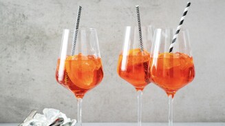 Drie glazen met Aperol Spritz, geserveerd met ijsblokjes en een rietje.