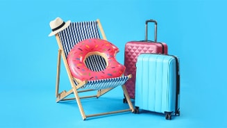 Strandstoel met strohoed, roze opblaasbare zwemband en twee koffers op blauwe achtergrond.
