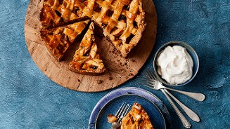 Zelfgemaakte appeltaart met ruitjespatroon op een houten plank, naast een schaaltje slagroom en een bord met een punt appeltaart en vork.