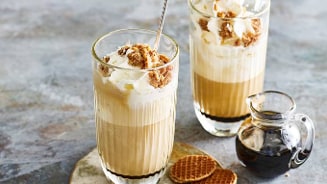 Twee glazen ijskoffie met slagroom en verkruimelde stroopwafels, naast een klein kannetje stroop en twee halve stroopwafels op een schotel.