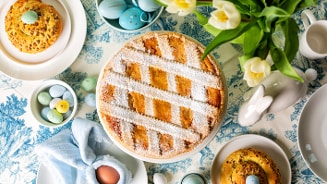 Paastafel met een grote traditionele taart met poedersuiker, decoratieve gekleurde eieren, kleine broodjes en een vaas met gele tulpen.