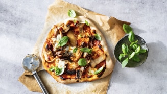 Zelfgemaakte pizza met champignons, kaas en verse basilicum op bakpapier, naast een pizzasnijder en een schaaltje met basilicumblaadjes.