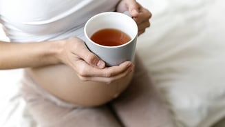 Zwangere vrouw met een kop thee in haar handen, zittend op een bed.
