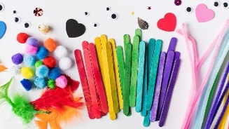 Knutselsupplies zoals gekleurde ijslollystokjes, pompoms, chenille draad, knutselveren, wiebelogen en foamvormen verspreid op een witte ondergrond