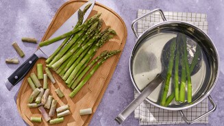 Verse groene asperges die worden voorbereid: sommige asperges zijn gesneden op een snijplank met een mes, terwijl enkele asperges in een pan met heet water liggen om te worden gekookt.