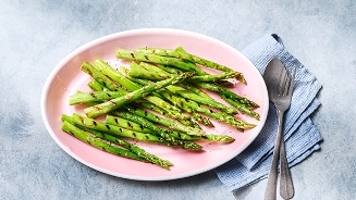 Gegrilde groene asperges op een roze bord met bestek en een servet.