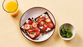 Twee crackers met cottage cheese, plakjes aardbei en verse basilicum op een bord, naast een glas sinaasappelsap en een schaaltje basilicum.