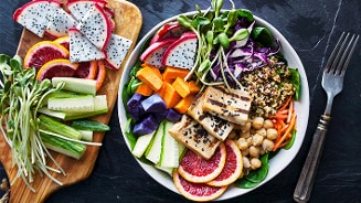 Vegetarische buddha bowl met verse groente, tofu, kikkererwten, quinoa, paarse zoete aardappel, rode kool, dragonfruit en plakjes bloedsinaasappel, geserveerd op een bord en een houten plank