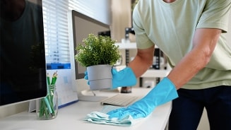 Persoon met blauwe schoonmaakhandschoenen die een bureau schoonmaakt en een plantenpot vasthoudt, naast een computer en ander kantoormateriaal.