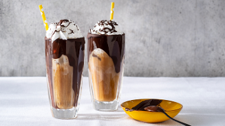 Twee glazen ijskoffie met slagroom en chocoladesaus, geserveerd met gele rietjes en een schaaltje met extra chocoladesaus.