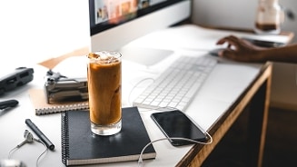Glas ijskoffie op een bureau naast een notitieboekje, oortjes en smartphone, met een computer op de achtergrond.