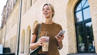Jonge vrouw met een schoudertas die buiten loopt, terwijl ze een smartphone vasthoudt en een beker ijskoffie drinkt.