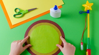 Kind knutselt een groene cirkel van gekleurd papier op tafel, omringd door schaar, lijm, gekleurd papier en een toverstaf