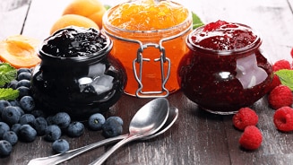 Potten met verschillende soorten jam omringd door vers fruit zoals bosbessen, frambozen en abrikozen op een houten tafel.