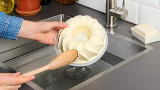Handen die een siliconen bakvorm schoonmaken met een houten lepel bij de gootsteen.