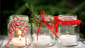 Kerstdecoratie met drie glazen potjes, elk met een brandend waxinelichtje, versierd met rood lint, dennentakjes en zuurstokjes