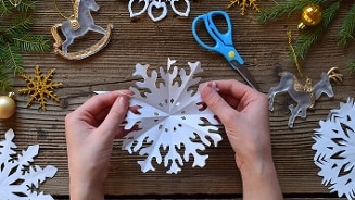 Handen maken een papieren sneeuwvlok als kerstversiering met schaar en kerstdecoraties op een houten tafel.
