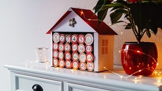 Adventskalender in de vorm van een huisje, versierd met kerstverlichting, op een dressoir naast een plant in een rode pot.