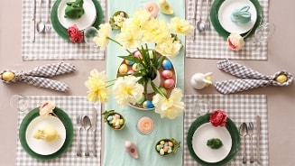 Paastafel met geel-witte bloemen in een vaas, kleurrijke paaseieren, servies op groene borden en decoratieve servetten, ideaal voor een feestelijk paasontbijt of paasbrunch.