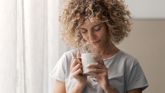 Persoon met krullend haar drinkt warme drank uit witte mok bij raam