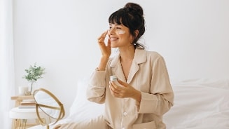 Vrouw in pyjama brengt crème aan op haar gezicht terwijl ze op bed zit, met make-up spiegel op de achtergrond