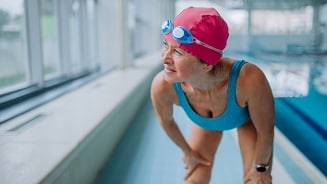 Vrouw in een blauw badpak en roze badmuts met zwembril, staand aan de rand van een binnenzwembad