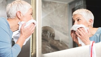 Persoon met grijs haar staat voor een spiegel en droogt het gezicht af met een handdoek in de badkamer
