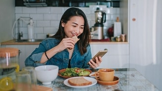 Vrouw eet ontbijt en kijkt op haar smartphone aan de keukentafel met brood, salade en koffie