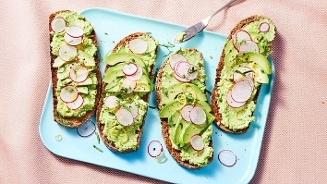 Vier sneetjes volkorenbrood belegd met groene spread, plakjes avocado en radijs op een lichtblauw bord