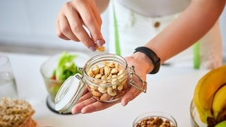 Hand pakt cashewnoten uit een glazen pot op een keukenblad, met fruit en salade op de achtergrond