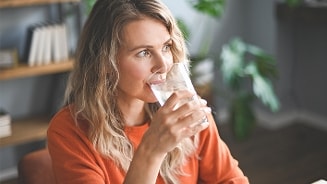 Vrouw in oranje trui drinkt een glas water in een woonkamer met planten op de achtergrond