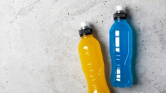 Twee plastic flessen sportdrank, één geel en één blauw, op een betonachtige achtergrond.