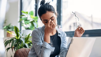 Vrouw zittend aan een bureau met een laptop, omringd door groene planten, houdt een bril vast in haar hand.
