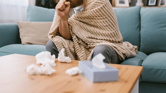 Iemand zit op een bank gewikkeld in een deken en houdt een zakdoek vast, met gebruikte tissues op de tafel.