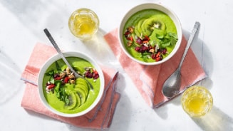 Twee kommen groene smoothie bowls met schijfjes kiwi, bessen en noten, op roze servetten met een glas geel sap.
