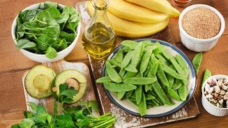 Verse en gezonde ingrediënten op een houten tafel, waaronder spinazie, avocado's, suikererwten, bananen, olijfolie, granen en gemengde bonen.