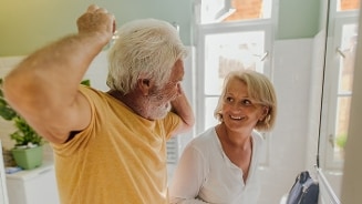 Oudere man en vrouw in een badkamer, waarbij de man voor de spiegel staat en zijn haar kamt