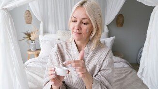 Vrouw in een kamer gehuld in een badjas, zittend op een bed met een potje crème in haar hand