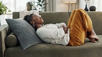 Vrouw in witte blouse en okergele broek liggend op een groene bank met een grijs kussen in een woonkamer