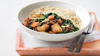 Kom met bruine rijst, linzen, tofu, wortel en spinazie geserveerd op een oranje plank met bestek ernaast