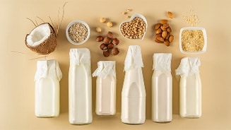 Verschillende soorten plantaardige melk in flessen, met kokosnoot, haver, hazelnoten, amandelen, rijst en kikkererwten op de achtergrond.