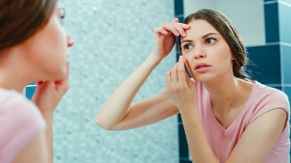 Persoon in een roze shirt die voor een spiegel staat en aan het gezicht huidverzorging toepast in de badkamer