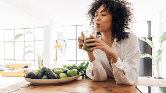 Persoon drinkt groene smoothie aan tafel met avocado's en limoenen in moderne keukenomgeving.