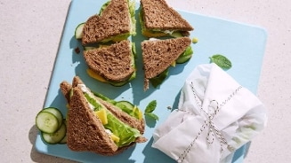 Volkoren sandwiches met kaas, sla en komkommer op een lichtblauwe snijplank, deels ingepakt in bakpapier